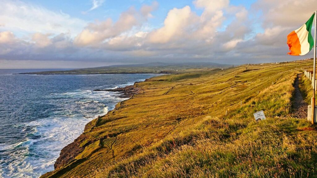 Panoramic views of the west coast of Ireland. In the foreground you can see the irish flag, green grass, cliffs, blossoms, in the background there is the sea with the coast and some waves.
