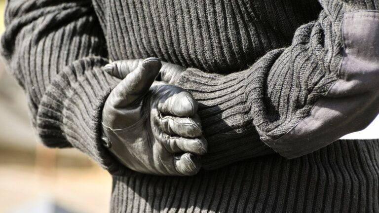 A man wearing a black jumper and gloves, styled on an Irish Republican paramilitary uniform, standing with his hands behind his back in a military pose